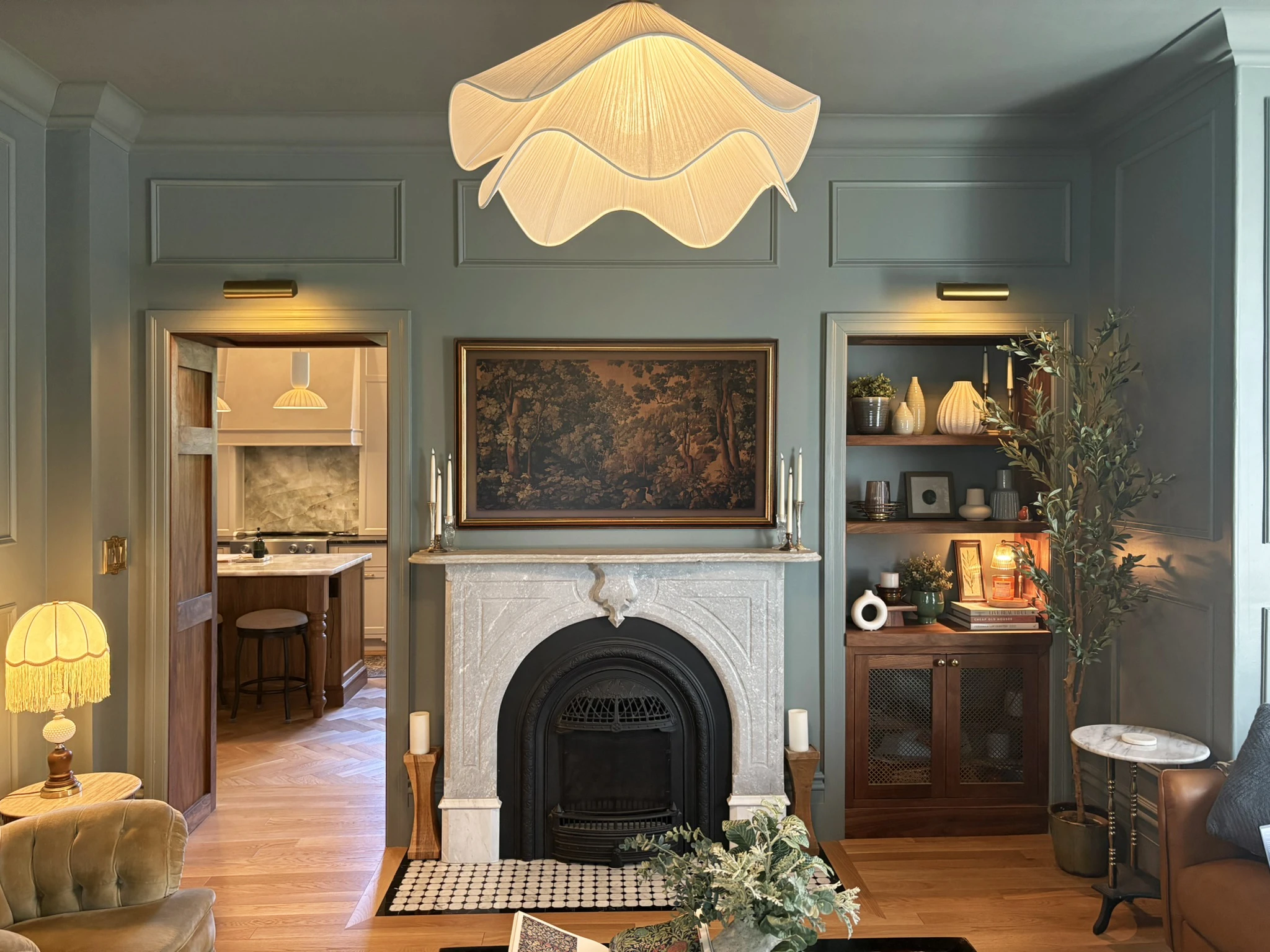Living room fireplace with layered lighting, paneled walls, and a view into the kitchen