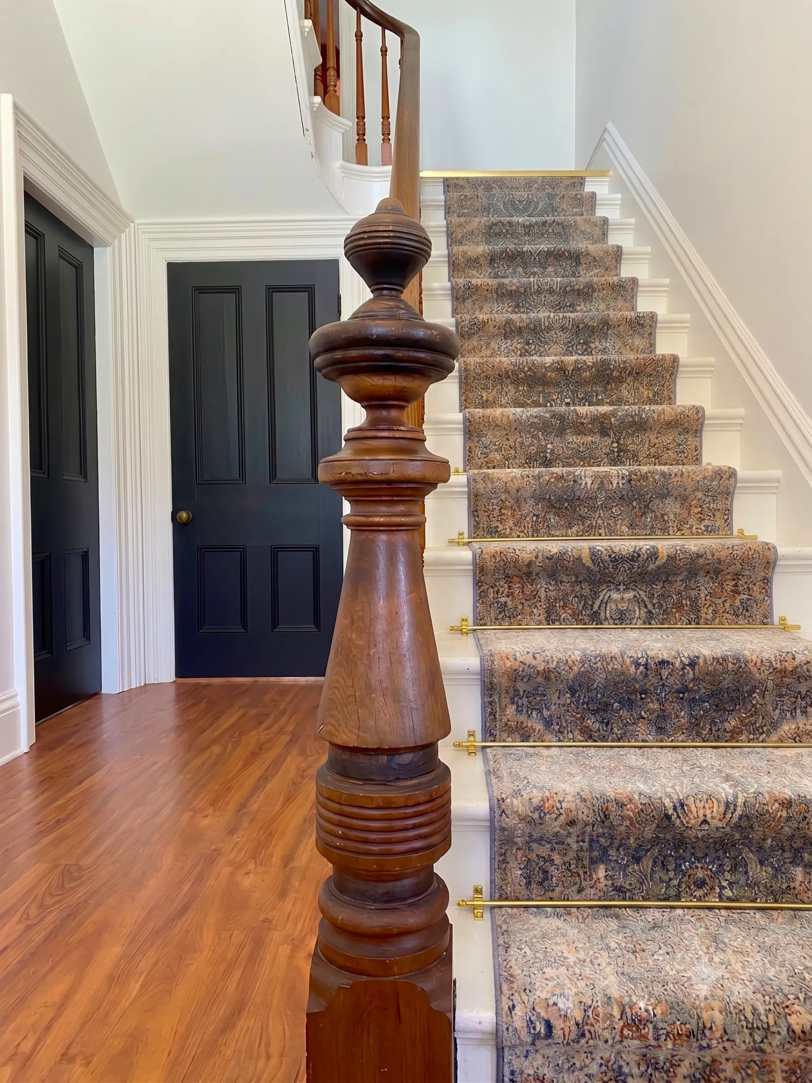 Hallway stair with patterned runner, brass stair rods, carved wood newel, and black doors